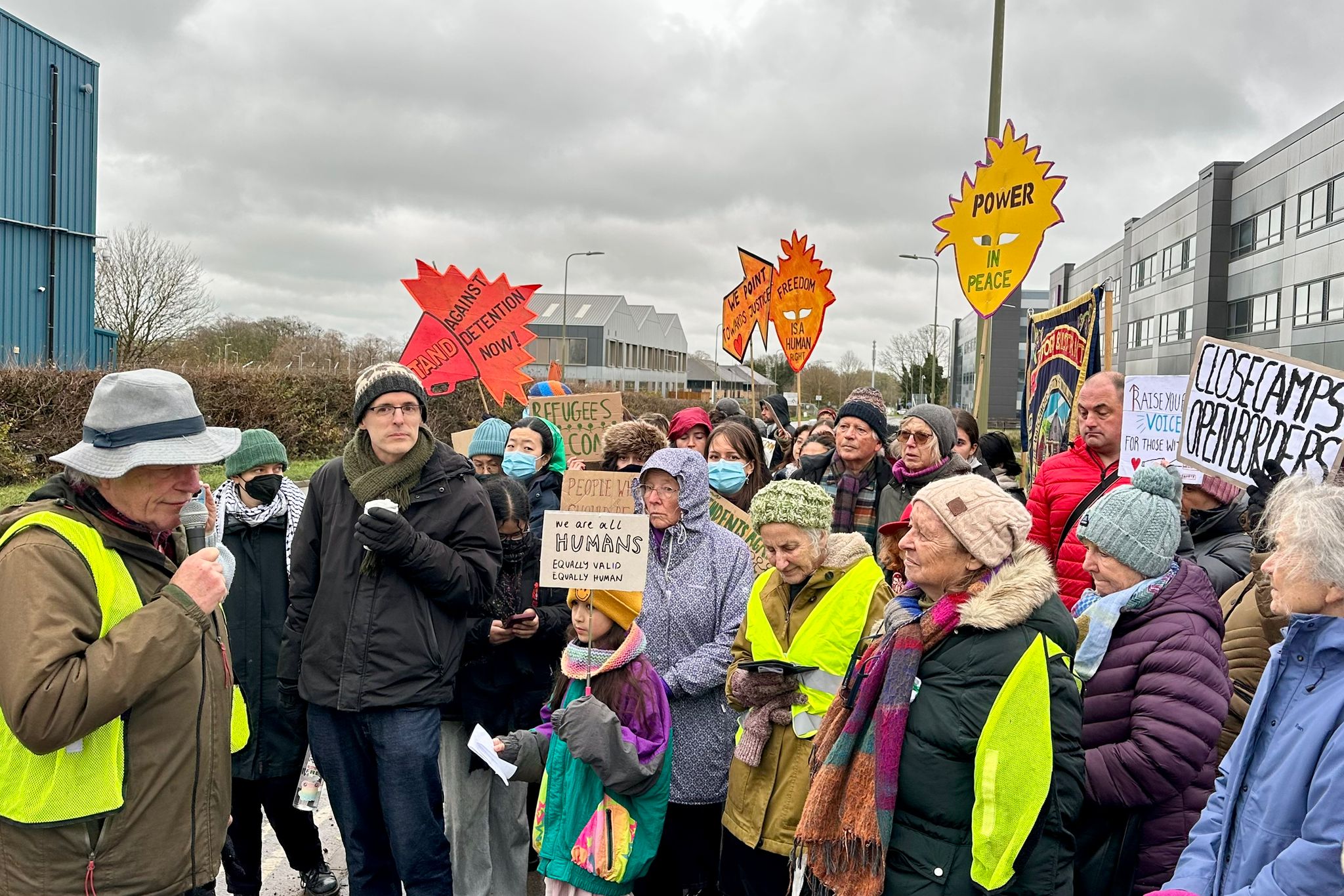 Roadside view with a sign pointing to “Ambulance Station” and “Campsfield House” on the left, while a large group of protesters with placards gathers on the pavement in the distance outside the Campsfield site under an overcast sky.