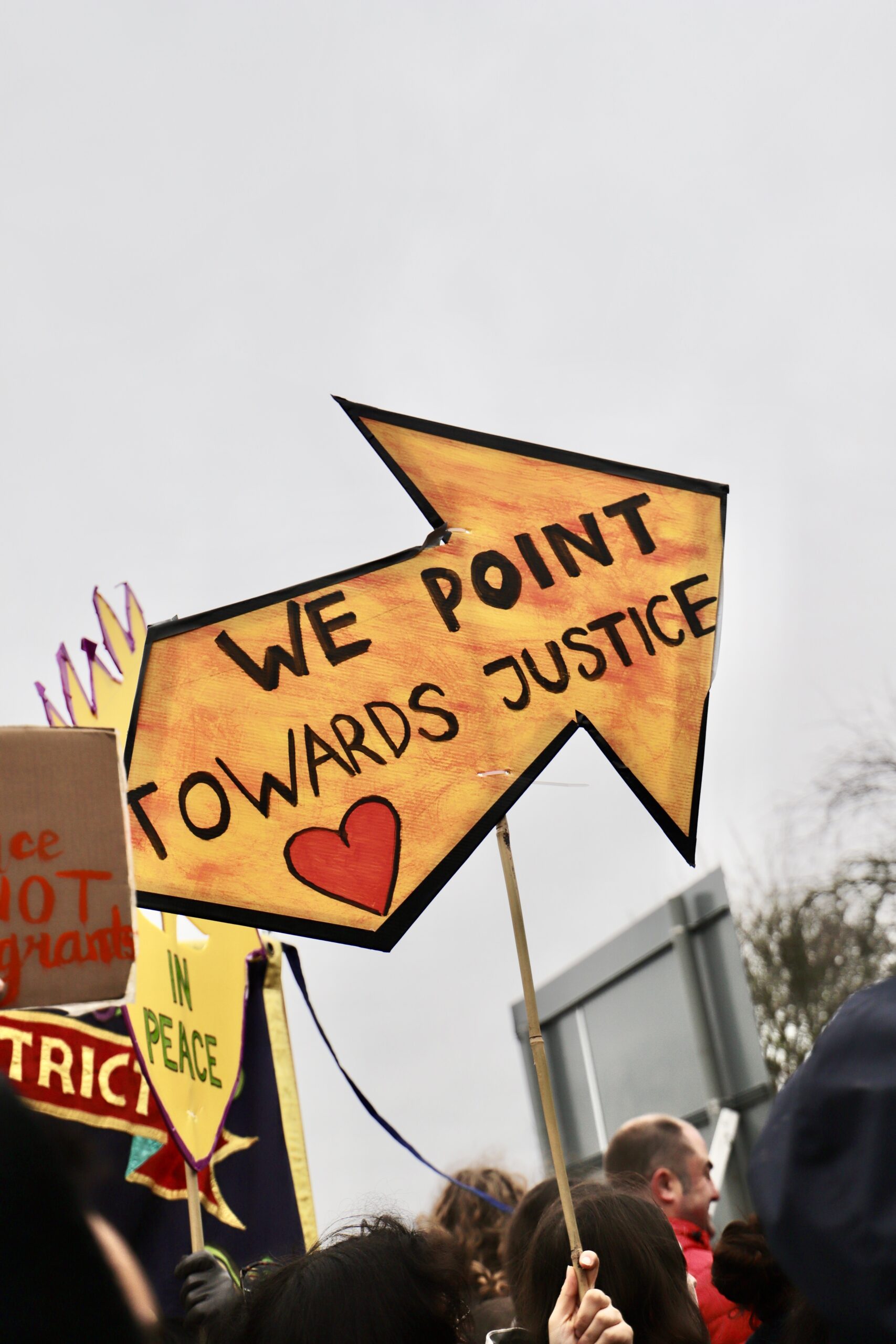 A large hand-painted yellow arrow-shaped protest sign reading “WE POINT TOWARDS JUSTICE” with a red heart, held above a crowd of demonstrators against a grey sky, with other colourful placards partially visible nearby.