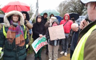 A group of protesters stand closely together in wet weather, listening to a speaker in a high-visibility vest, with several people holding handmade “Keep Campsfield Closed” signs and umbrellas in the background.
