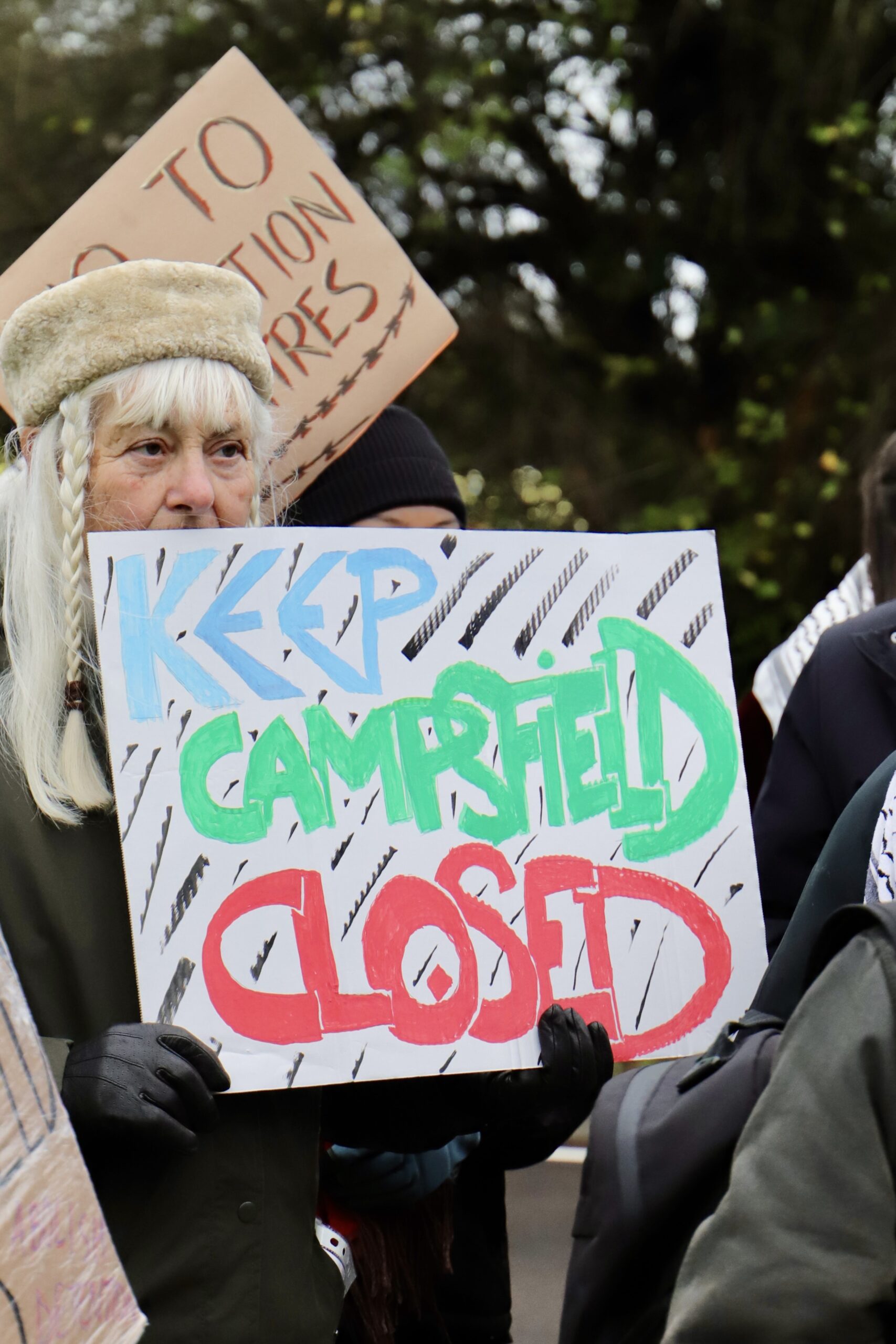 A protester holds a handmade placard reading “Keep Campsfield Closed” in bright green, blue and red lettering, surrounded by other demonstrators and banners at an outdoor protest.