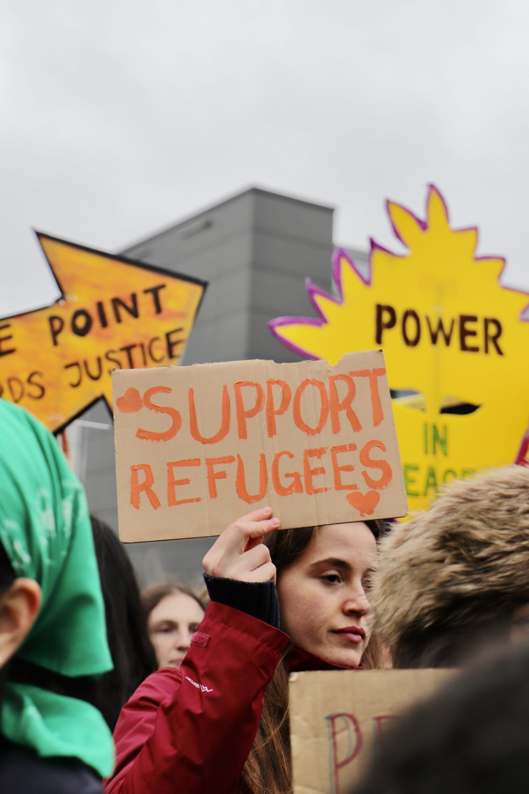 A young protester in a red jacket holds up a cardboard sign reading “SUPPORT REFUGEES” with small hearts, standing among a crowd of demonstrators with brightly coloured protest signs and buildings in the background under a grey sky.