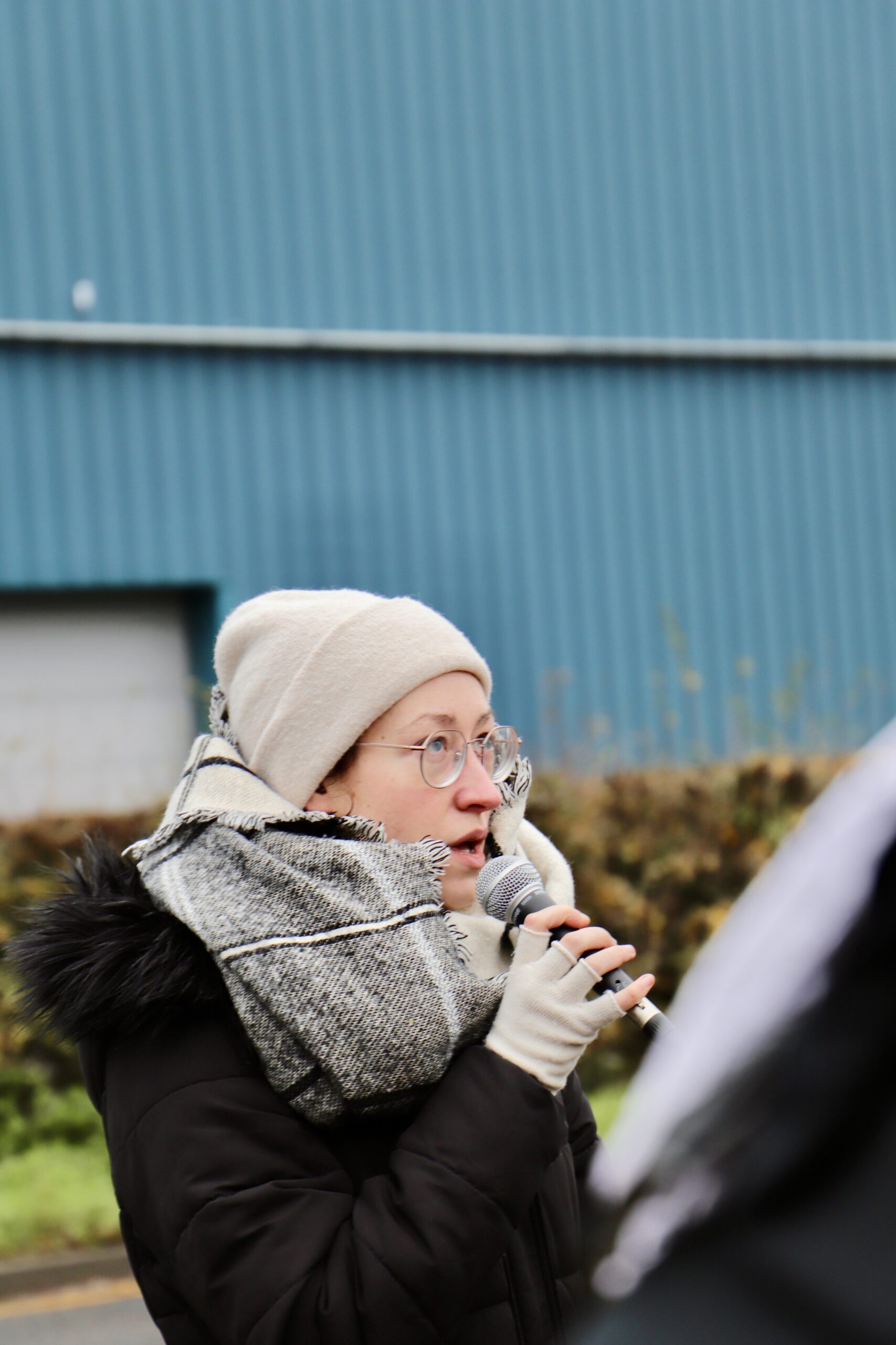 A speaker wearing a beige beanie, glasses and a thick scarf holds a microphone and addresses the crowd outside Campsfield, with blue industrial buildings and winter hedges in the background.