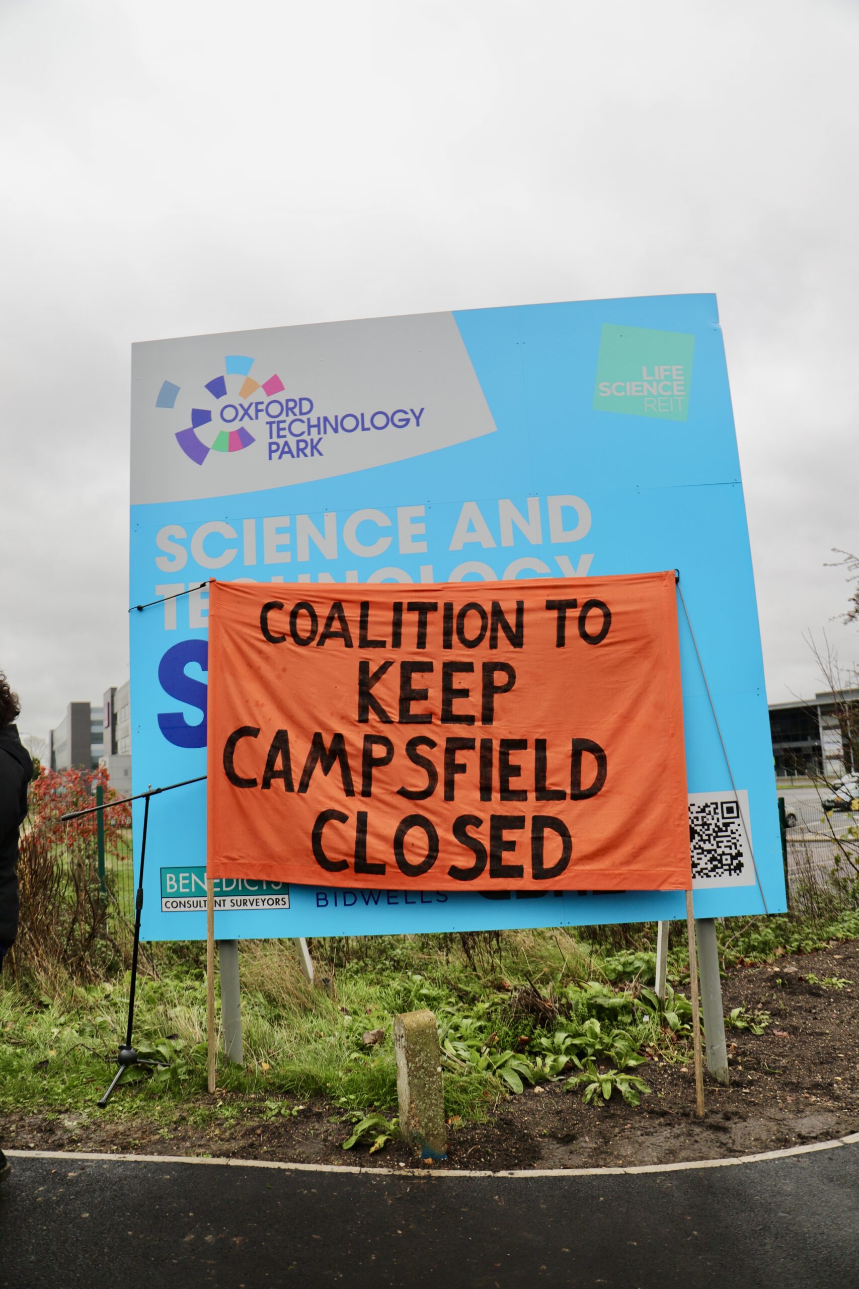 An orange banner reading “COALITION TO KEEP CAMPSFIELD CLOSED” is tied across a large blue Oxford Technology Park sign that says “Science and Technology”, beside a roadside verge under an overcast sky.