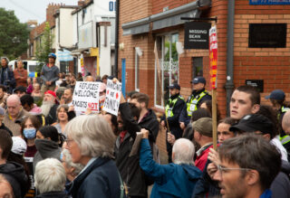 A large crowd of people holding placards. One placard says "Refugees Welcome! Racists not!"