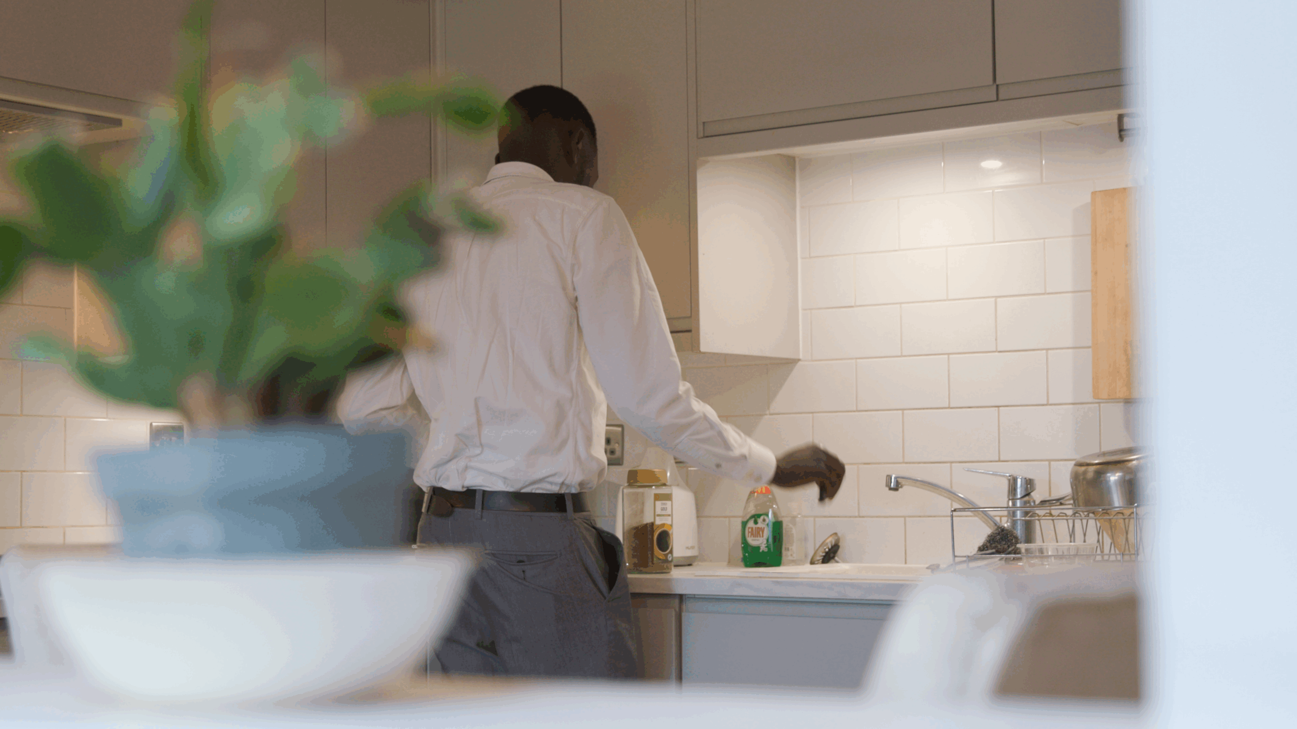 A person wearing a white shirt stands at a kitchen counter near a sink, with washing-up liquid and jars visible; a green plant is blurred in the foreground.