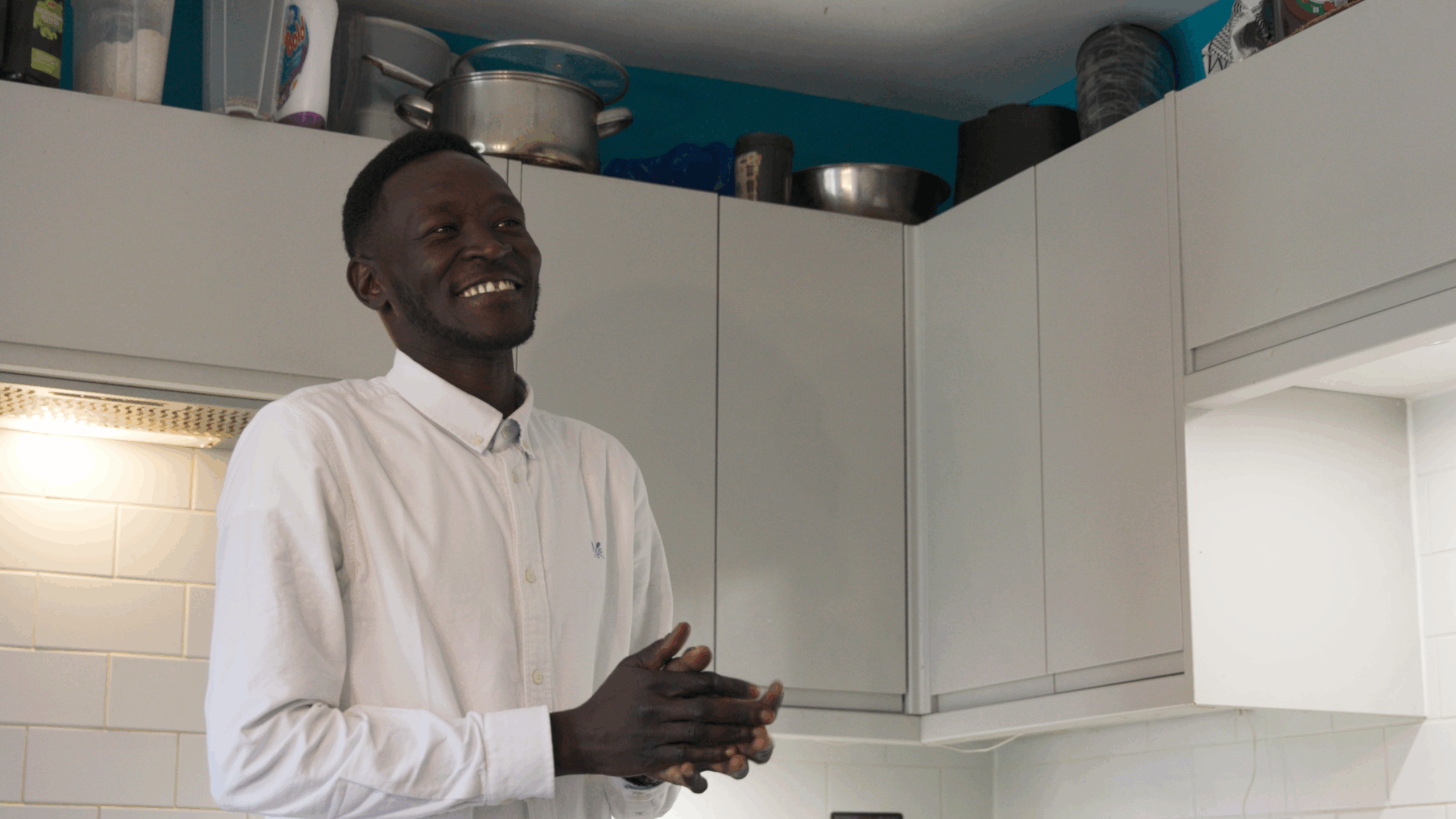 A man in a white shirt stands smiling in a kitchen, hands together as if mid-conversation. Pots and bowls are stacked on the cupboards above him.