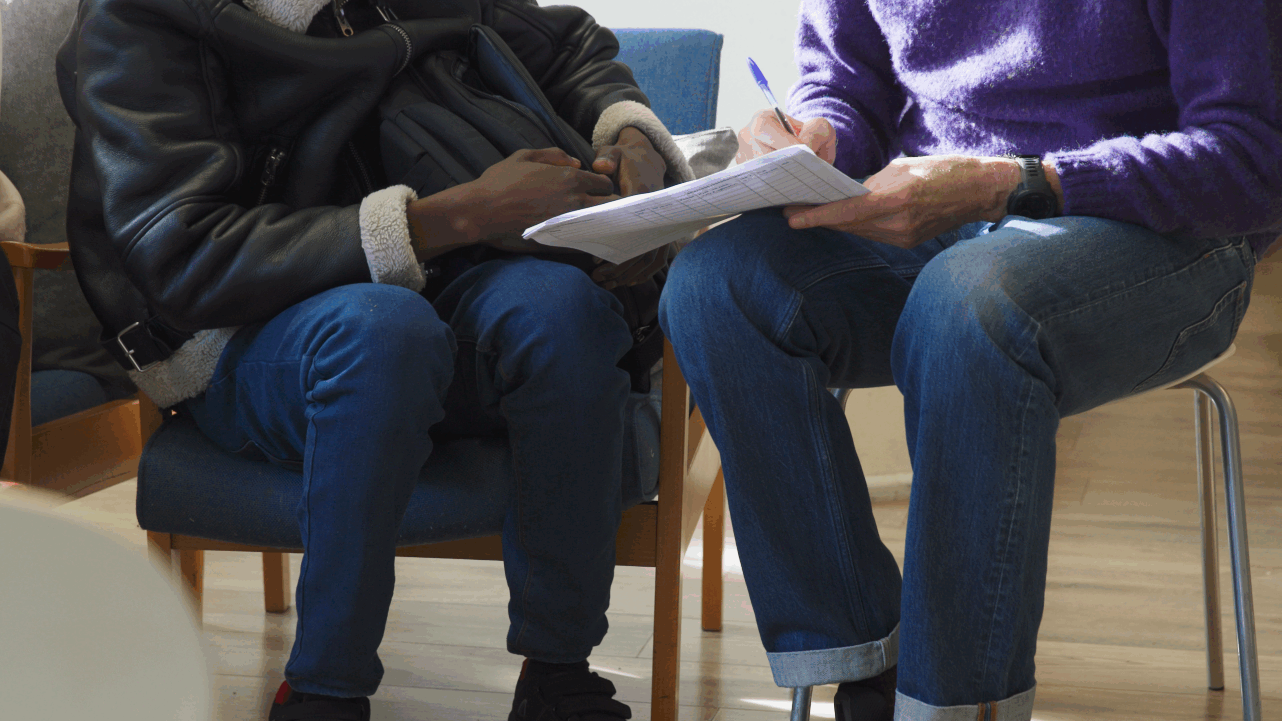 Close-up of two people sitting side by side, one taking notes on a clipboard while the other holds a backpack. Only their lower bodies are visible, suggesting a meeting or interview.